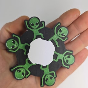 May include: A circular black and white alien-themed decoration. Six green aliens with large eyes are arranged around a white center. The aliens have their arms raised in a playful pose. The decoration is held in a hand.