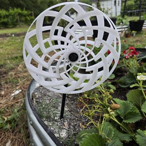 May include: A white, spherical garden ornament with a geometric, openwork design. The ornament is mounted on a black metal stake and is set in a garden bed with strawberry plants and red flowers.