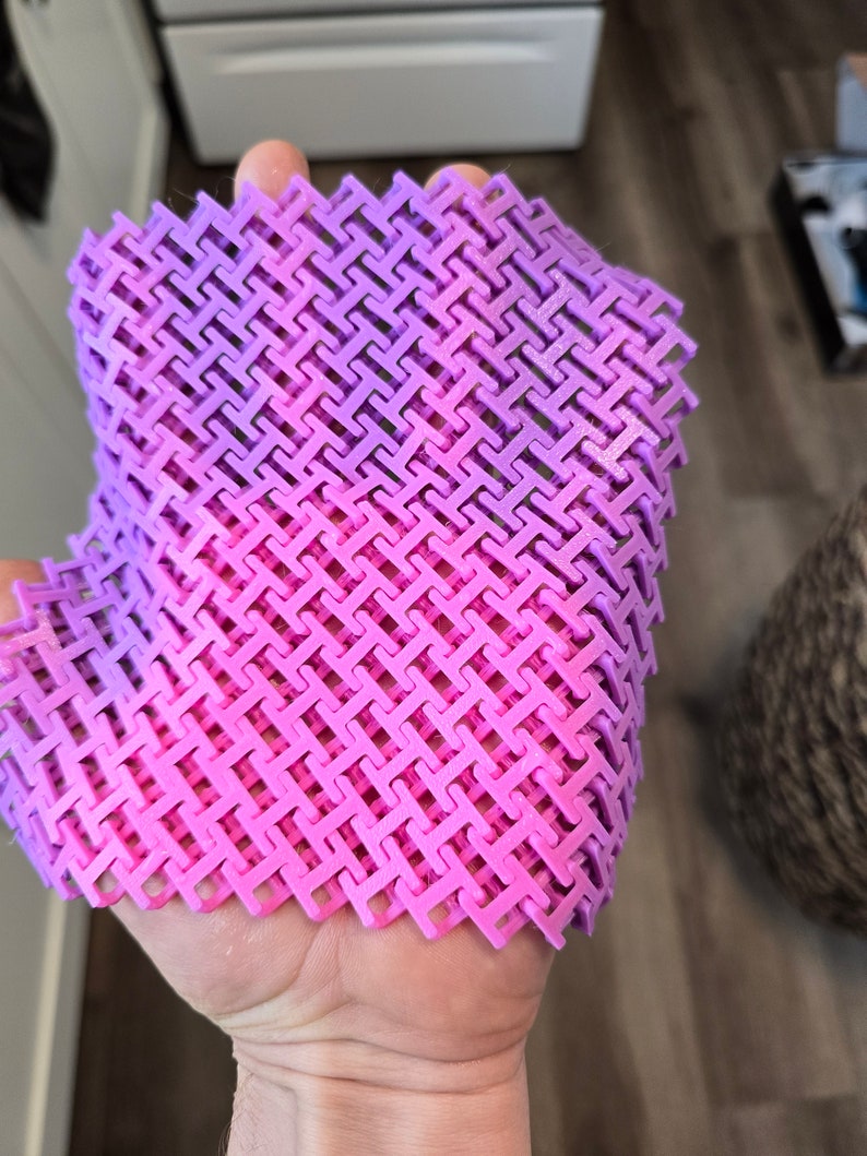 May include: A flexible, 3D-printed object with a woven pattern. The object transitions from pink to purple, held in a hand. The intricate design suggests a unique texture and form, possibly a decorative or functional item.
