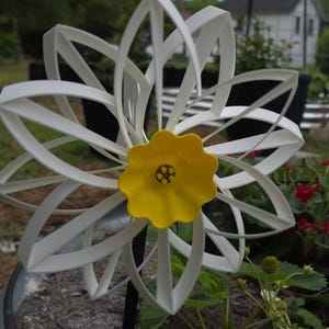 May include: A garden spinner featuring a white, multi-layered flower design with elongated, looped petals. The center is a bright yellow, scalloped-edge disc with a small, dark brown circular detail in the middle, all mounted on a thin black rod.