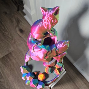 May include: A multi-colored 3D printed dice tower with a cat figurine on top. The tower has a circular base with a groove for holding dice. The cat figurine is sitting on a platform with a hole in the center. The tower is surrounded by a variety of multi-colored dice.