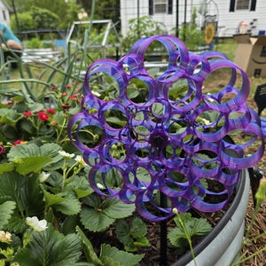 May include: A purple and blue garden ornament with a circular design, resembling a cluster of bubbles, is placed in a garden bed with strawberry plants. The ornament is made of a metallic material and is mounted on a black stake.