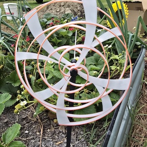 May include: A decorative garden pinwheel with a spiral design. The pinwheel features white blades and orange trim, mounted on a black pole. It is set in a garden bed with green plants and flowers.