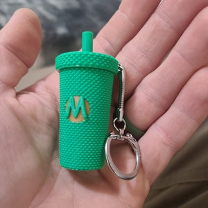 May include: A green plastic keychain shaped like a cup with a straw and a brown "M" logo. The keychain has a silver metal ring.