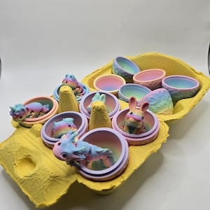 May include: A yellow egg carton with pastel rainbow colored eggs. Each egg contains a different pastel rainbow colored animal figurine. The animals include a unicorn, a bunny, a dragon, and a cat.