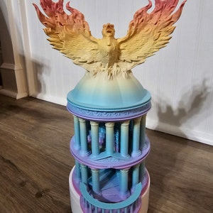 May include: A 3D printed figurine of a phoenix rising from a multi-colored, tiered temple. The phoenix is painted in shades of yellow, orange, and red, while the temple is painted in shades of blue, purple, and pink.