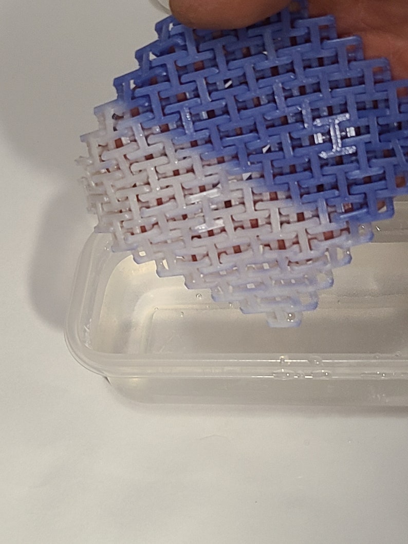 May include: A blue and white plastic coaster with a woven design. The coaster is held above a clear plastic container. The coaster's design is a repeating pattern of interlocking shapes.