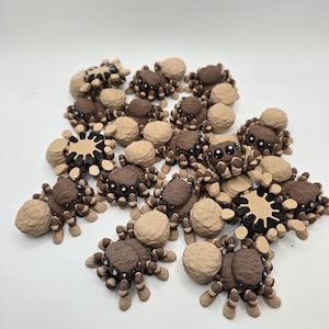 May include: A collection of small, detailed spider figurines in various shades of brown and tan. The spiders have rounded bodies and multiple legs, with some featuring intricate patterns. The figurines are arranged on a white surface.