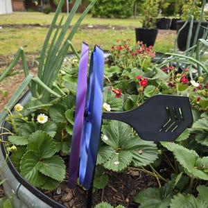 May include: A dual-layered pinwheel featuring a purple front blade and a blue back blade, attached to a black plastic stake with a horizontal fin. The pinwheel is positioned vertically within a garden bed containing green strawberry plants.