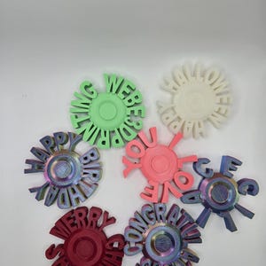 May include: A collection of colorful, circular decorative objects with various celebratory messages. The objects are in shades of green, white, pink, red, and iridescent colors. Messages include "Happy Birthday", "Merry Christmas", and "Congratulations".