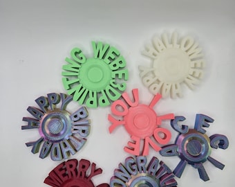 May include: A collection of colorful, circular decorative objects with various celebratory messages. The objects are in shades of green, white, pink, red, and iridescent colors. Messages include "Happy Birthday", "Merry Christmas", and "Congratulations".