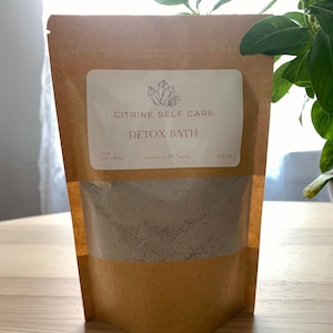 May include: A brown paper bag of Citrine Self Care Detox Bath. The bag has a clear window showing the gray powder inside. A white label with the brand name and product name is affixed to the front. The bag is on a wooden surface.