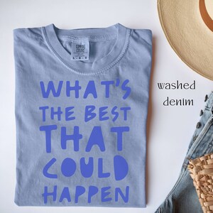 May include: A light blue t-shirt with the text "WHAT'S THE BEST THAT COULD HAPPEN" printed in blue. The shirt is folded and laid flat on a white surface.