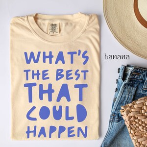 May include: A light beige t-shirt with the text "WHAT'S THE BEST THAT COULD HAPPEN" printed in blue. The shirt is laid flat on a white surface with a straw hat and blue jeans in the background.