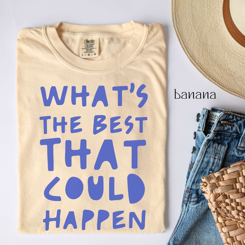 May include: A light beige t-shirt with the text "WHAT'S THE BEST THAT COULD HAPPEN" printed in blue. The shirt is laid flat on a white surface with a straw hat and blue jeans in the background.