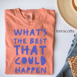 May include: A terracotta coloured t-shirt with the text "What's the best that could happen" printed in purple.