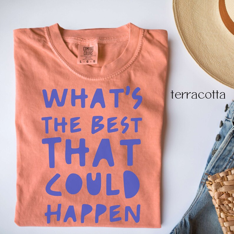 May include: A terracotta coloured t-shirt with the text "What's the best that could happen" printed in purple.