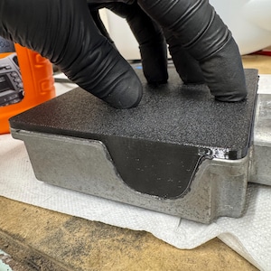 May include: A black textured rectangular gasket is being pressed onto a silver metal component. The component has a recessed area filled with the same black material as the gasket. The image is taken from a close-up perspective.