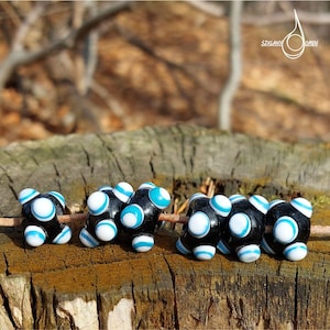 May include: Black glass beads with white and blue accents are displayed on a wooden surface. The beads have a unique design, with white and blue stripes and circles. The beads are arranged in a row, showcasing their intricate details.