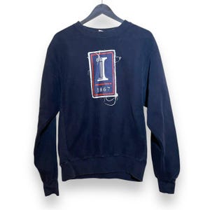 Op de afbeelding: Een marineblauw sweatshirt met lange mouwen en een rechthoekige patch met een wit zuilicoon, het getal 1867 en rood-witte randen. De patch heeft zichtbare gerafelde randen en losse draden, en het kledingstuk is voorzien van een ronde hals.