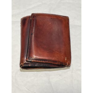 May include: A tri-fold wallet crafted from distressed brown leather. The wallet features visible wear along the edges and a smooth, polished surface with natural grain variations. The construction includes stitched seams and a rectangular shape.
