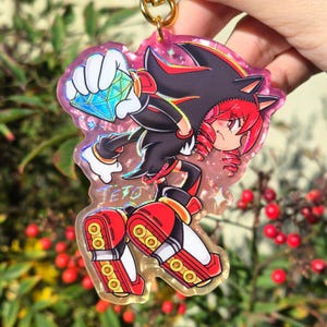 May include: A vibrant acrylic keychain depicting a cartoon character with black and red fur, clutching a blue gem. The keychain has a gold-coloured ring and the name "TETO" is printed on the bottom. The background is a pinkish hue with a glitter effect.