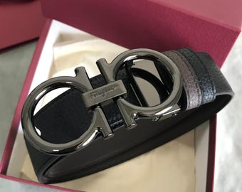 Può includere: Una cintura in pelle nera testurizzata con fibbia Gancini color canna di fucile e logo Ferragamo inciso sulla barra centrale. La cintura è arrotolata all'interno di una scatola bordeaux con rivestimento interno bianco dal design strutturato.