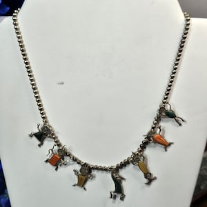 May include: A silver beaded necklace featuring multiple dangling charms shaped like stylized figures. Each figure is inlaid with colorful stone accents in shades of green, orange, and yellow. The necklace has a polished metallic finish.