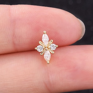 Puede incluir: Un pendiente de botón dorado con cuatro pétalos y piedras de circonita cúbica transparentes y brillantes. El pendiente presenta una piedra central en forma de marquesa y tres piedras redondas más pequeñas. El pendiente se sostiene entre dos dedos.