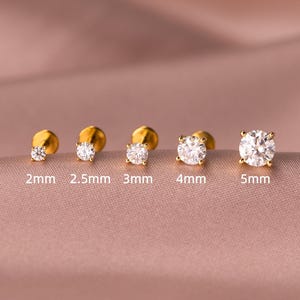 Puede incluir: Una colección de pendientes de botón dorados con piedras de circonita cúbica redondas y transparentes. Las tallas varían de 2 mm a 5 mm, con cada tamaño de pendiente etiquetado. Presentados sobre un fondo rosa suave.