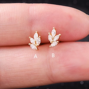 May include: Two gold-colored leaf-shaped earrings, each adorned with multiple clear, marquise-cut gemstones. The earrings are displayed on a light-toned background, with the letter "A" next to one and "B" next to the other.