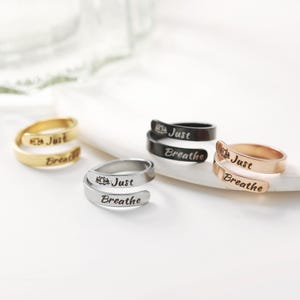 May include: Four silver rings with the words "Just Breathe" and a lotus flower design. The rings are in different colors: gold, black, silver, and rose gold.
