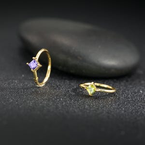 May include: Two gold rings with square gemstones. One ring features a purple gemstone, and the other has a green gemstone. The rings are set against a dark background with a large, dark gray stone.