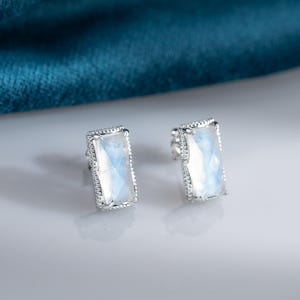 Puede incluir: Un par de pendientes de botón rectangulares con un engaste plateado. Cada pendiente presenta una gran piedra preciosa rectangular iridiscente de color azul claro y blanco. Los bordes de los pendientes están detallados con pequeñas cuentas plateadas.