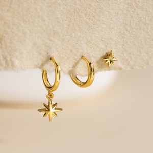 May include: A set of gold earrings. The set includes two small hoop earrings and a single stud earring. One hoop earring has a star charm. The earrings are displayed on a cream-coloured surface.