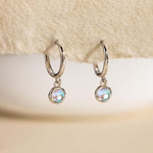 May include: Two silver hoop earrings, each with a small, iridescent, round charm. The earrings are shown against a neutral background. The charms have a shimmering, pearlescent effect, reflecting a range of colours. The hoops are a simple, elegant design.