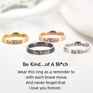 Puede incluir: Cuatro anillos en oro, oro rosa, negro y plata, cada uno grabado con "...of a bitch" y "Be Kind". Los anillos se muestran sobre una superficie blanca. El texto "Be Kind...of A Bi*ch" está debajo.
