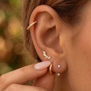 May include: A gold ear cuff with small clear stones, a gold earring with four marquise-cut clear stones arranged in a curve, a small gold hoop earring, and two tiny gold stud earrings with single clear stones worn in a single ear.