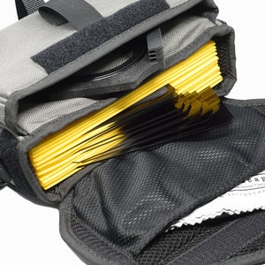 Könnte beinhalten: Eine graue Kameratasche aus Canvas mit einem schwarzen Riemen und einer Reißverschlussöffnung. Die Tasche ist offen und enthält einen Stapel gelber Filter und ein schwarzes Reinigungstuch.
