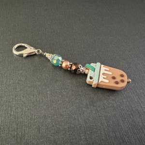 May include: A brown and white charm with a brown and white milk tea design, with a silver charm attached to a silver chain with blue and brown beads.