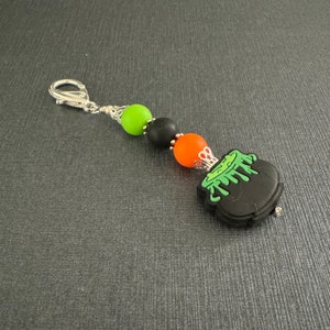 May include: A black cauldron charm with green slime dripping down the sides. The charm is attached to a silver chain with green, black, and orange beads.