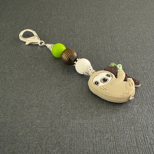 May include: A silver key chain with green, brown, and white beads and a brown sloth charm. The sloth is holding a branch and has a cute face.