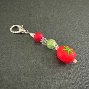 May include: A red tomato charm with green leaves, attached to a silver chain with red and green beads.