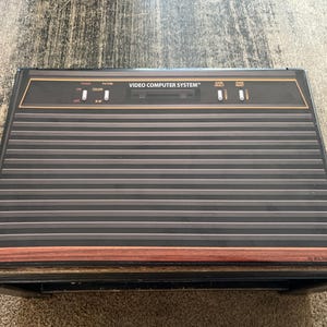 Puede incluir: Una consola de videojuegos negra y marrón con el texto "VIDEO COMPUTER SYSTEM" en la parte delantera. La consola tiene una serie de líneas horizontales y una moldura de madera.