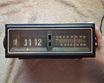 May include: A black General Electric flip clock radio featuring a digital-style flip display showing 11:12 PM, an analogue AM/FM radio tuner with a red needle, and a vertical slider switch for alarm settings on the front panel.