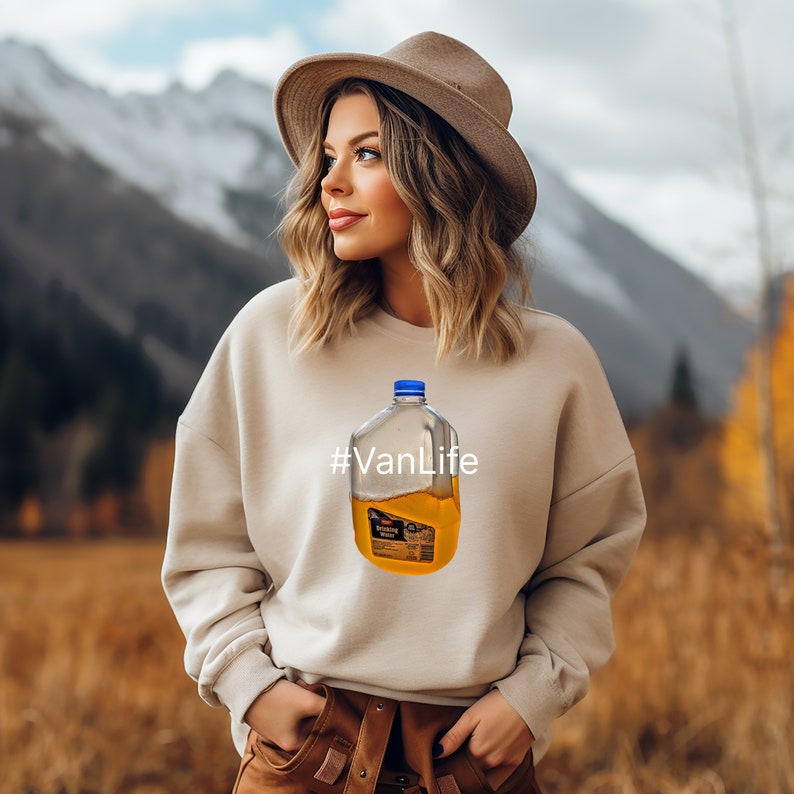 May include: A woman wearing a beige sweatshirt with a clear plastic jug of orange liquid printed on it. The jug has a blue lid and the text "#VanLife" is printed below it. She is wearing a brown hat and looking to the left. The background is a blurry image of a mountain range.