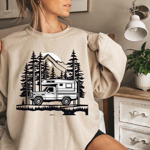 May include: Beige sweatshirt featuring a black and white graphic of a camper truck in a forest scene with mountains and a sun. The design includes silhouettes of pine trees and a detailed depiction of the vehicle.