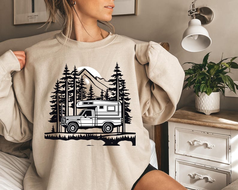 May include: Beige sweatshirt featuring a black and white graphic of a camper truck in a forest scene with mountains and a sun. The design includes silhouettes of pine trees and a detailed depiction of the vehicle.