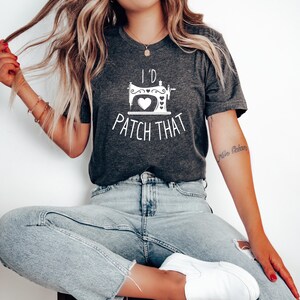 Puede incluir: Camiseta gris oscuro con el texto blanco "I'd Patch That". El diseño presenta una máquina de coser con un corazón y elementos decorativos. La camiseta está hecha de un material suave y es adecuada para uso casual.
