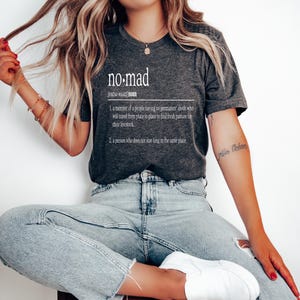May include: A dark gray t-shirt with a white graphic design that reads "no-mad" and defines the word as a person who does not stay long in the same place.